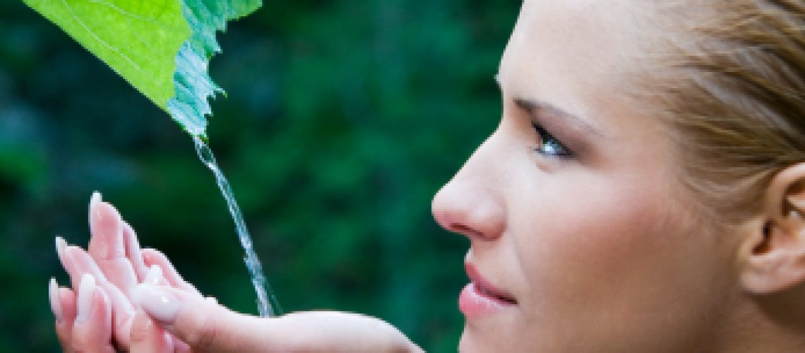 Beautiful young woman refreshing herself with clear water from a leaf in the nature. Symbol of purity and nature harmony